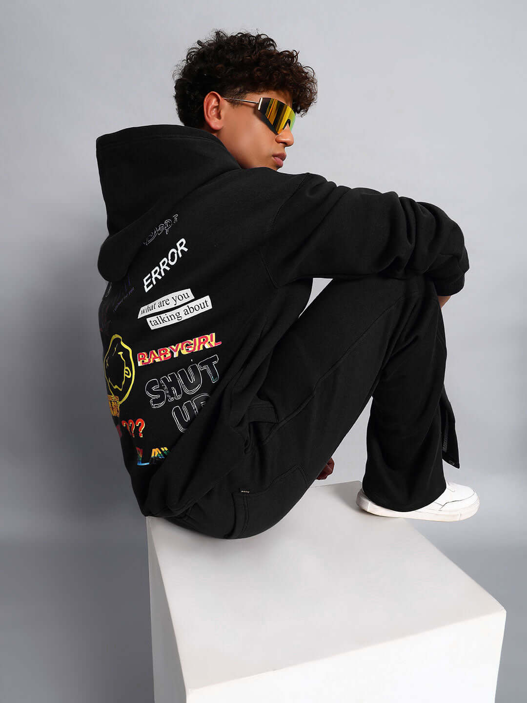 Model wearing WEARDUDS SUPREME FLEECE HOODIE BLACK featuring graphic patches, seated on white pedestal.