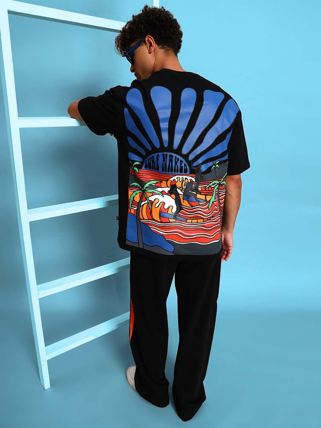 Back of man wearing SURF NAKED OVER-SIZED T-SHIRT BLACK with vibrant design, standing by a shelf against blue background.