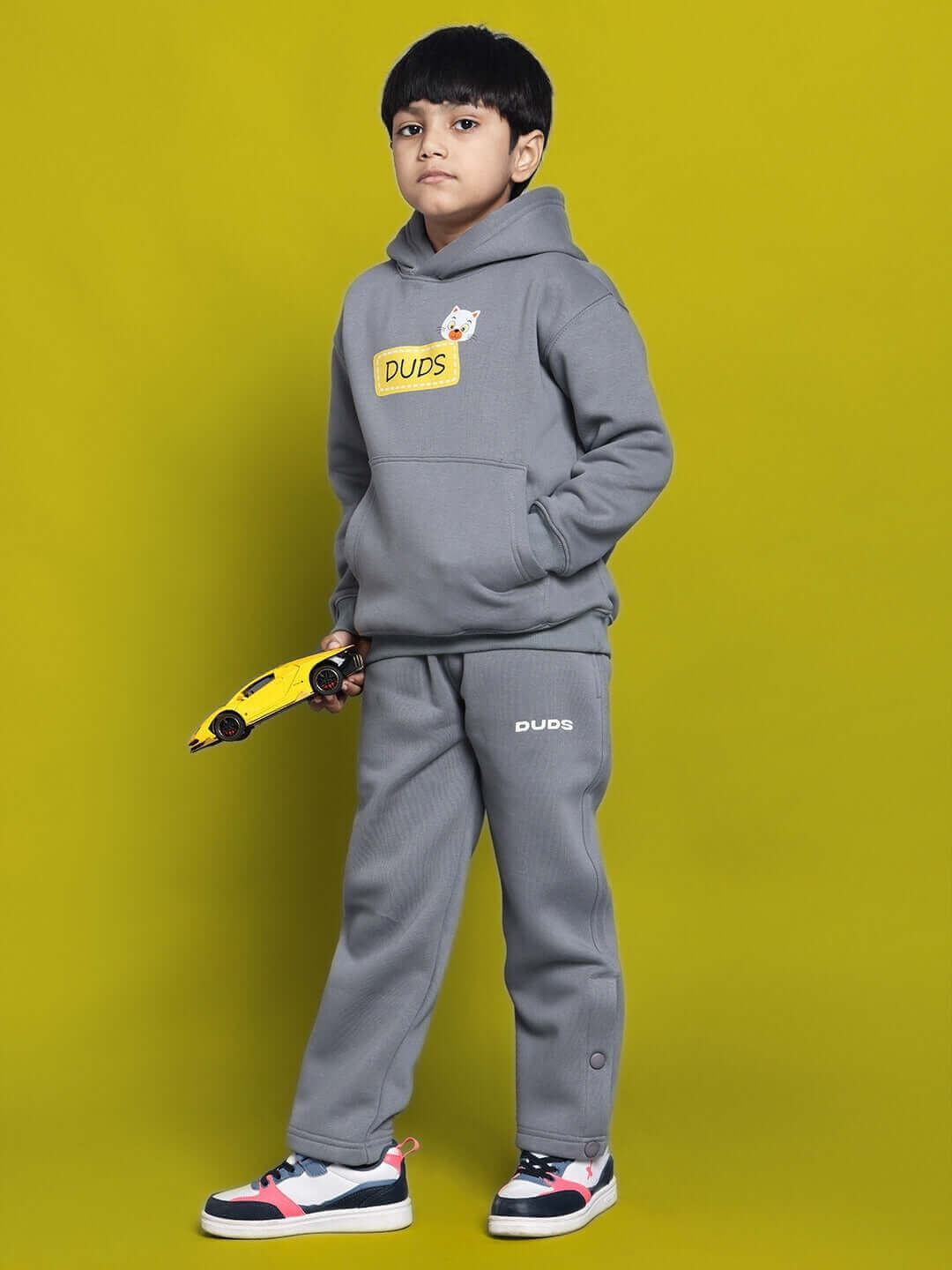 Child wearing grey Tabby fleece pant and hoodie set, holding a toy car, with a yellow backdrop.