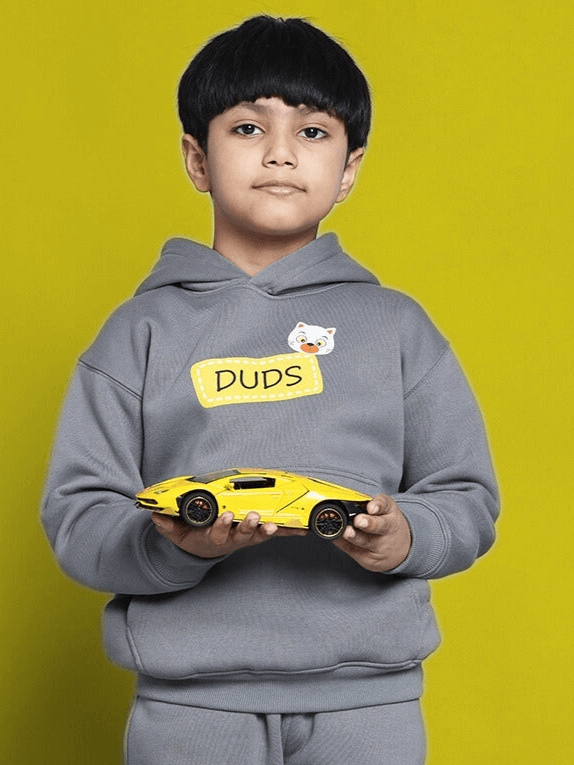 Child in TABBY OVERSIZED FLEECE HOODIE FOR BOYS GIRLS GREY, holding yellow toy car against yellow background.