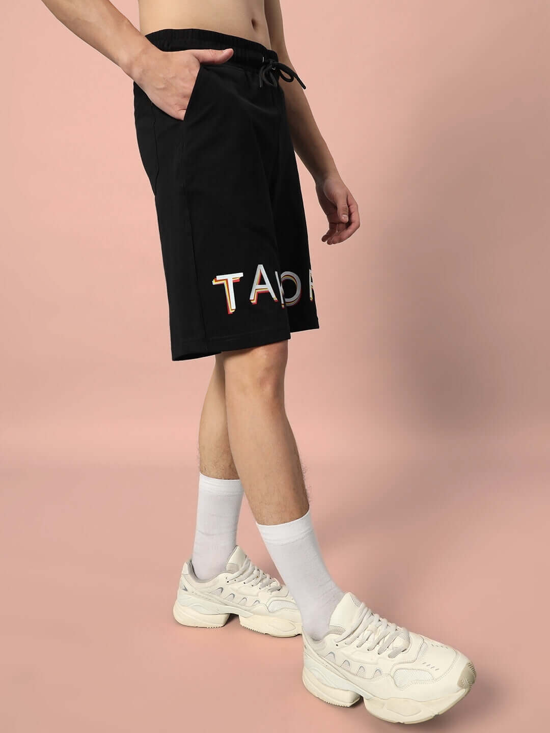 Man wearing TAKE OFF regular fit black shorts with embroidered logo, paired with white sneakers, against a peach background.