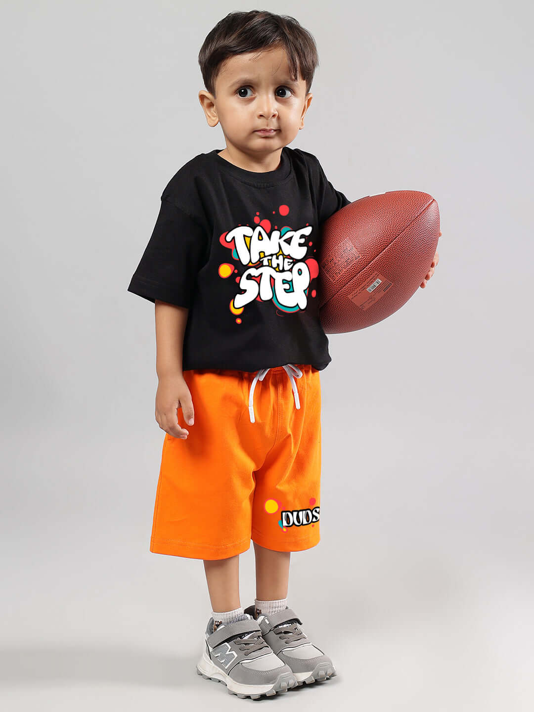 Young child in black 'Take the Step' shirt and orange DUDS shorts, holding a football, representing customizable kids' attire.
