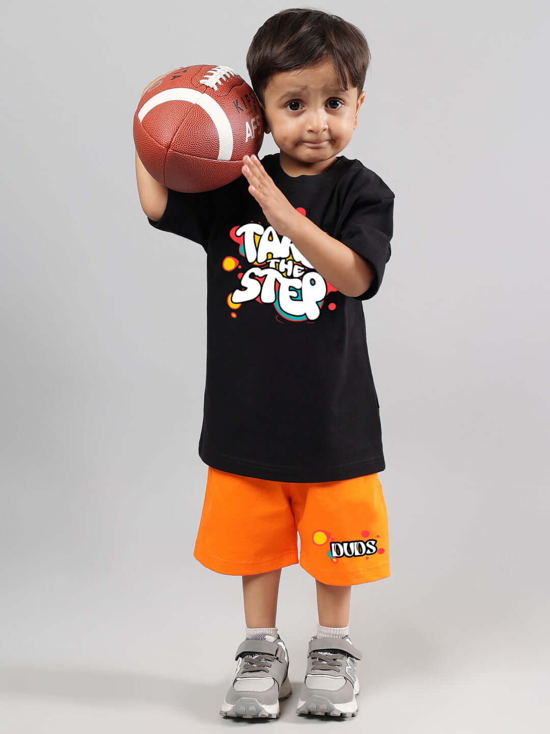 Child wearing a 'Take the Step' black t-shirt, holding a football, orange shorts with DUDS logo, perfect for boys and girls fashion.