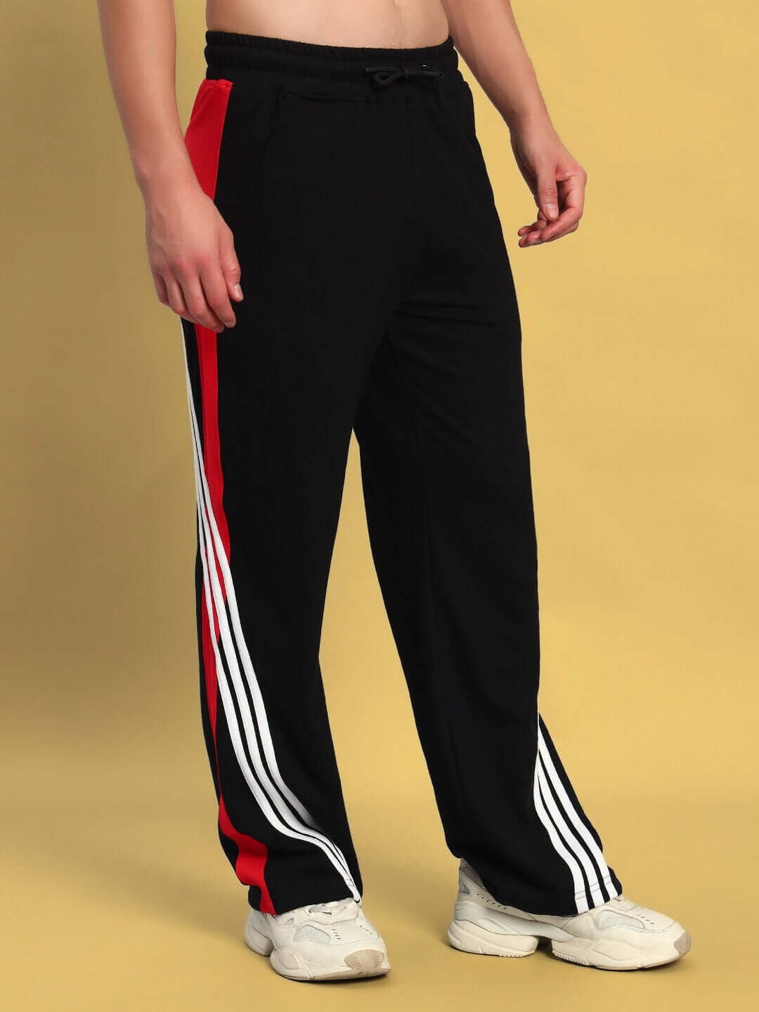 Model wearing black-red TANGLE relaxed-fit joggers by DUDS with white sneakers, featuring side stripes, on a yellow background.