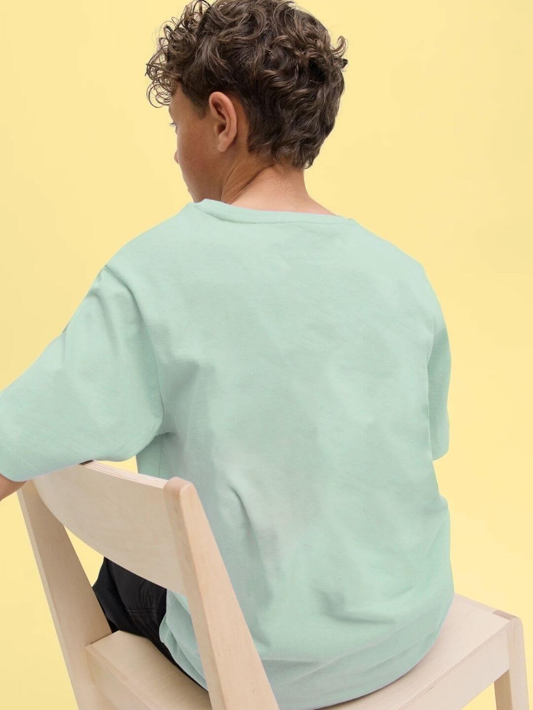 Child wearing TARGET PRINTED T-SHIRT FOR BOYS AND GIRLS LIGHT GREEN sitting on chair, back view in bright yellow background.