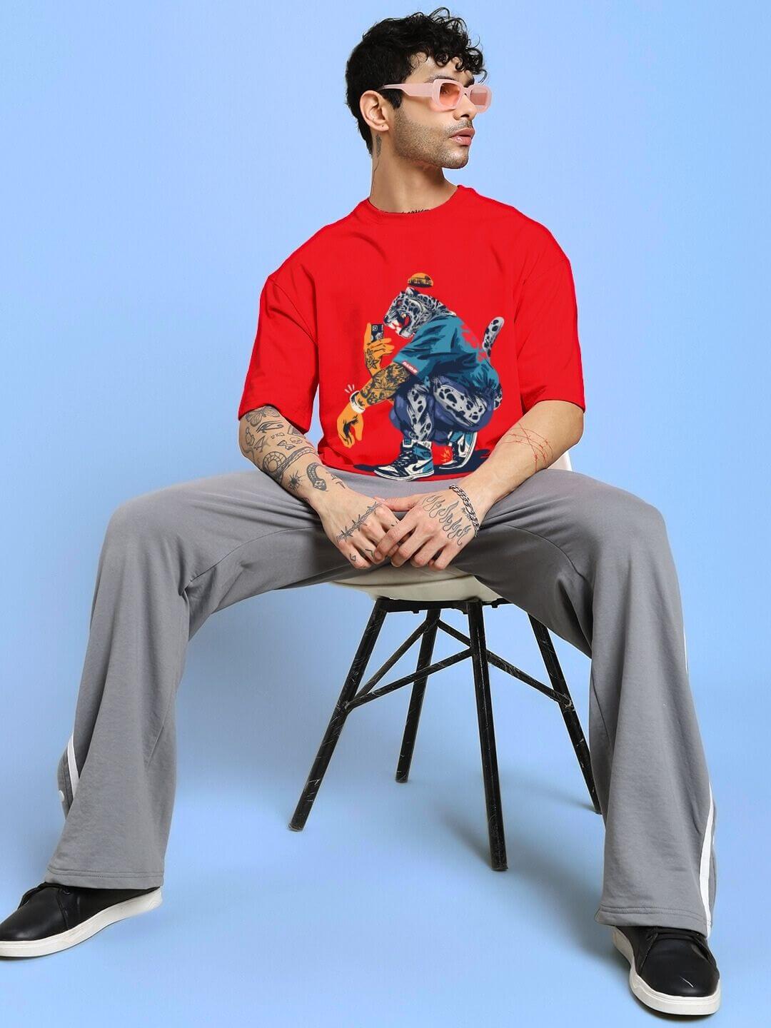 Man wearing TEKKEN KING T3 OVER-SIZED T-SHIRT RED, seated stylishly showcasing bold graphic and vibrant color on light blue background.