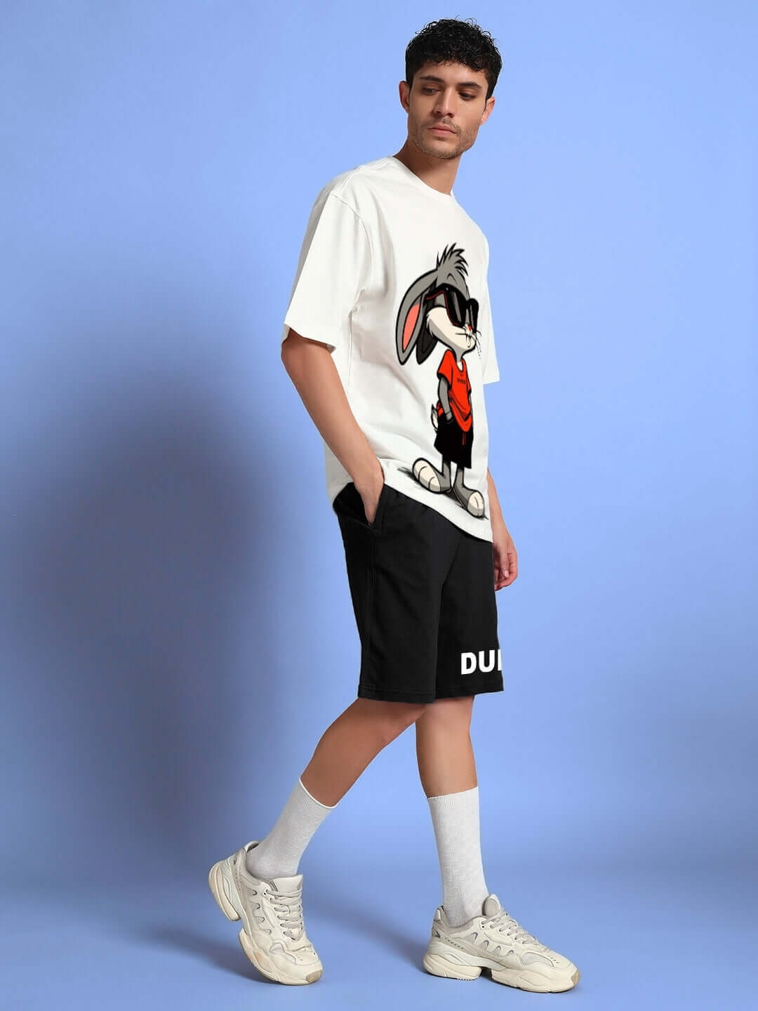 Man wearing Think Big Bunny Toons Co-ord Set in off-white and black, featuring a cartoon print, styled with sneakers against a blue background.