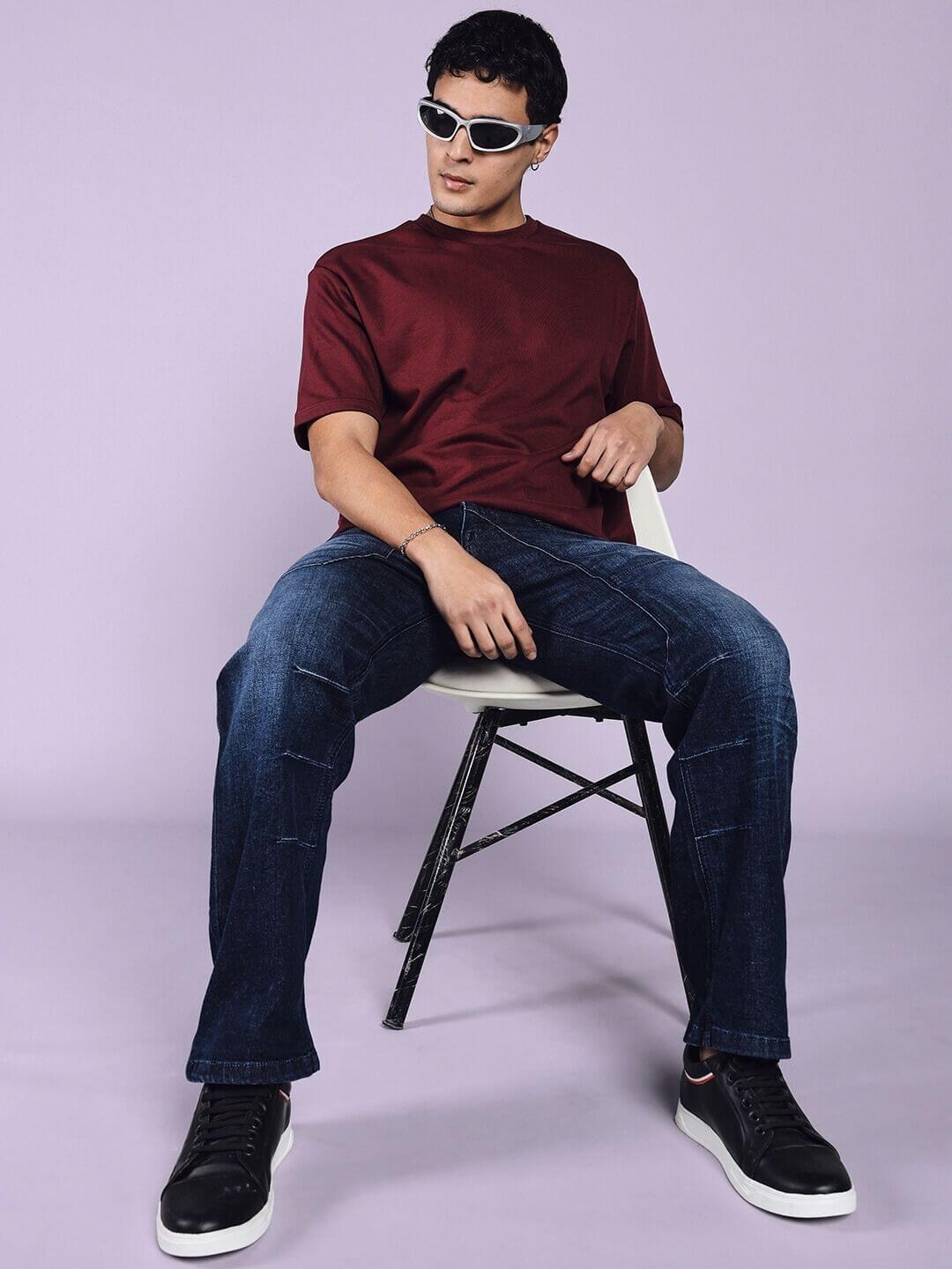 Model wearing Thrifty Regular Fit Denim Jeans in dark blue, paired with a maroon t-shirt and black sneakers, sitting on a chair.