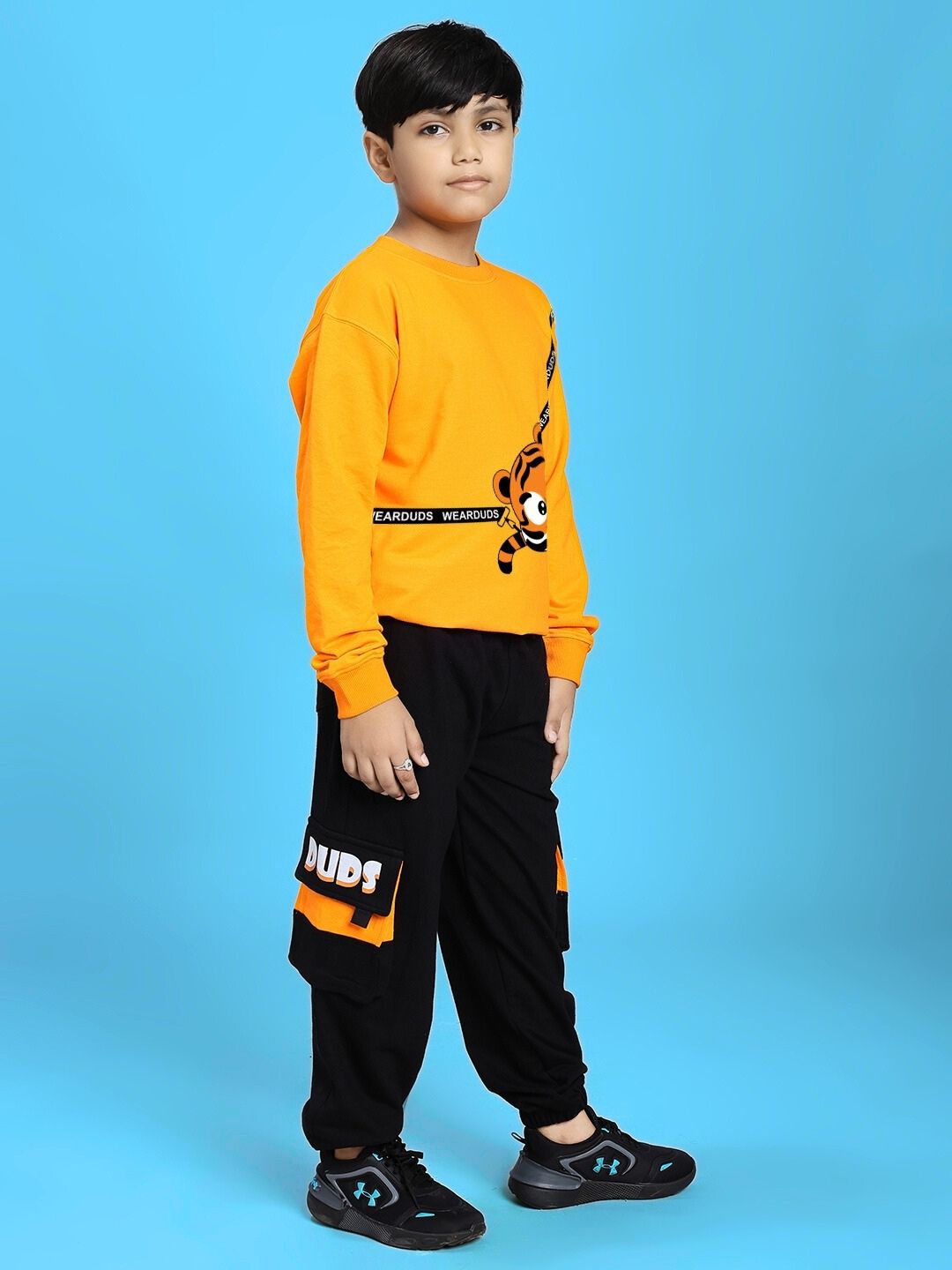 Child wearing orange sweatshirt and black joggers, posing against blue background