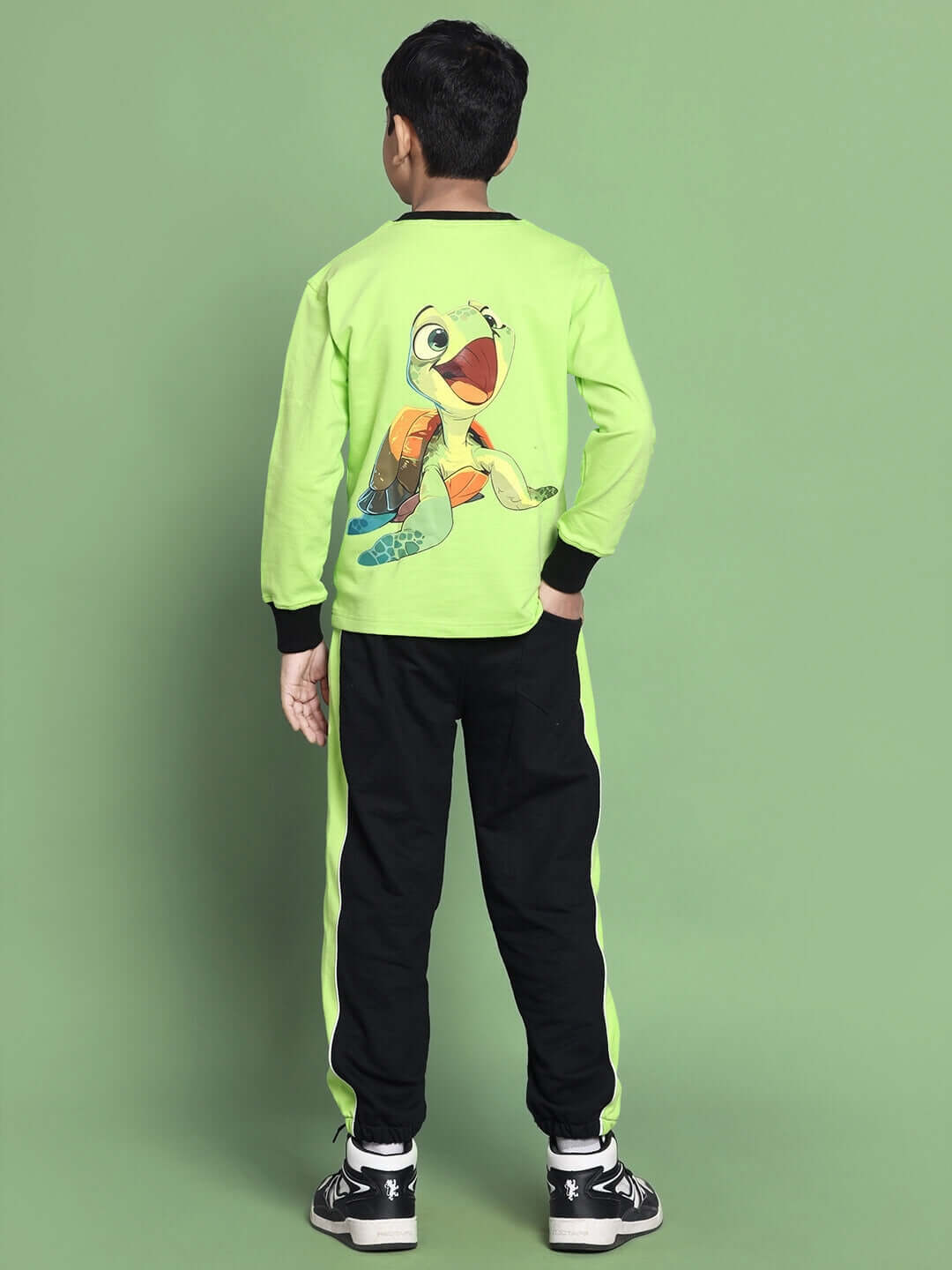 Boy wearing TILLY OVERSIZED CO-ORD SET FOR BOYS GIRLS NEON GREEN with cartoon print, black pants, and sneakers on green background.
