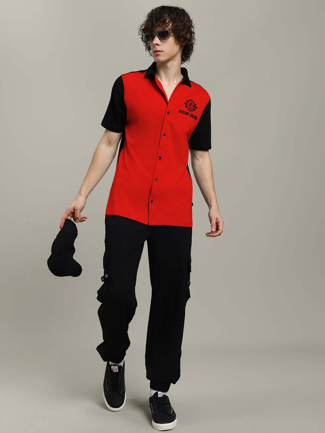 Man wearing TINGE regular fit red-black shirt, black pants, and sunglasses, holding cap. Made to order by DUDS tailors.