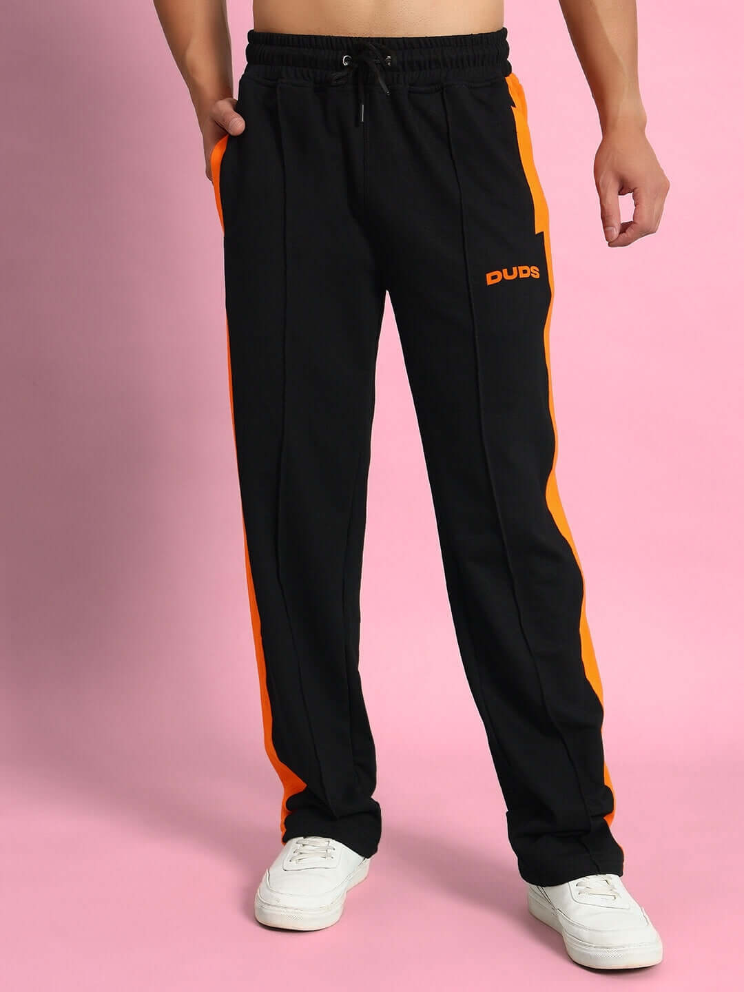 Black joggers with orange contrast panel, elastic waistband, and drawstring, styled on a person wearing white sneakers, pink background.