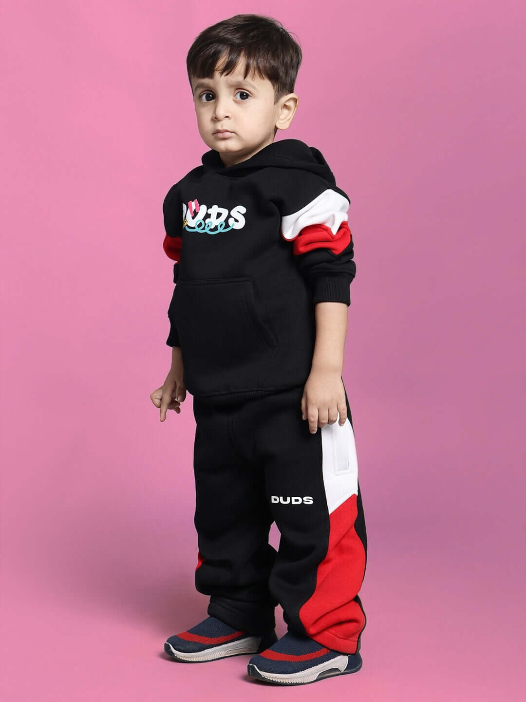 Child wearing black beanie fleece jogger outfit with red and white accents, standing against a pink background.
