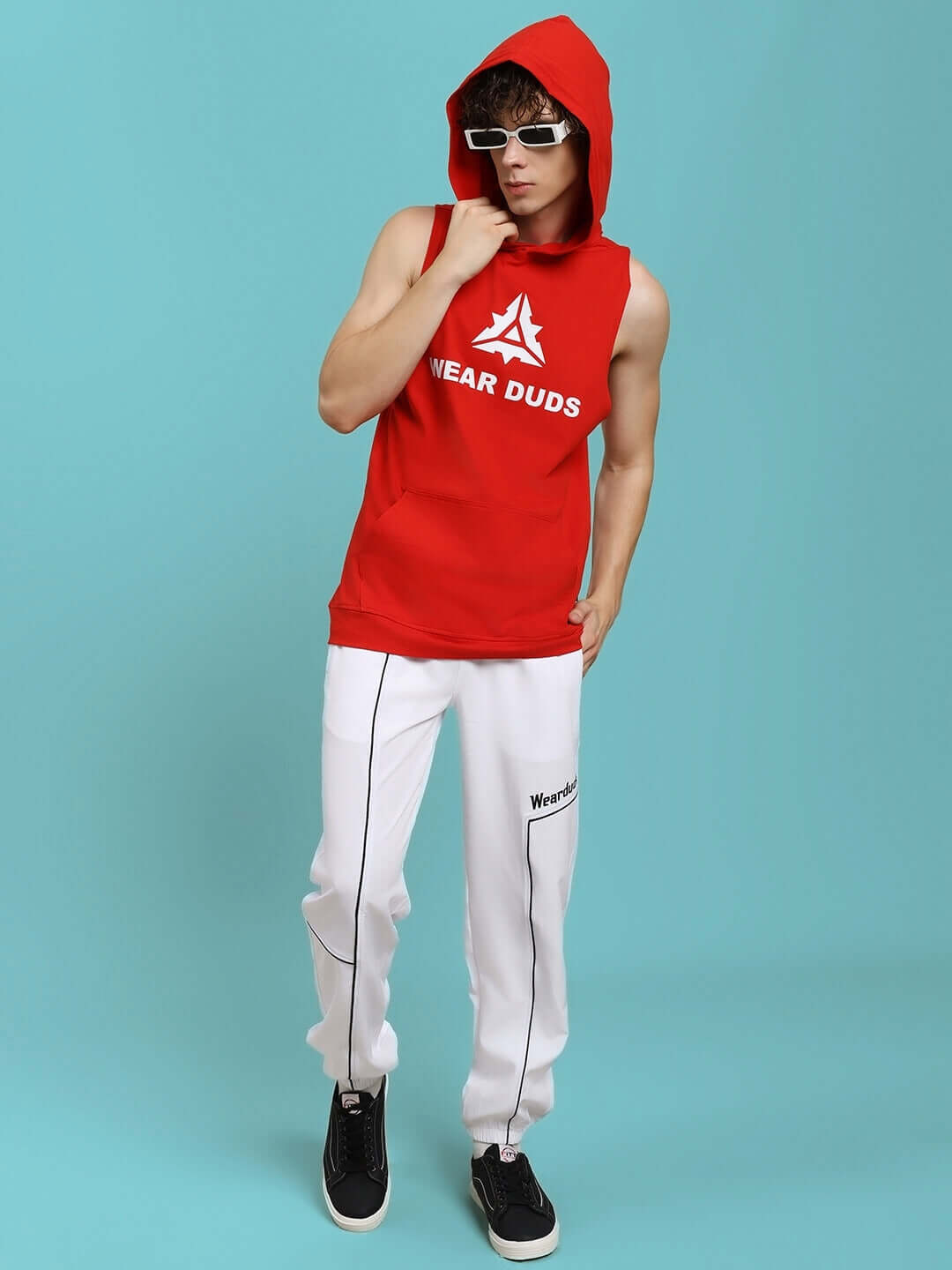 Man wearing TOKYO RELAXED FIT JOGGERS WHITE with red sleeveless hoodie and sunglasses, standing in studio.