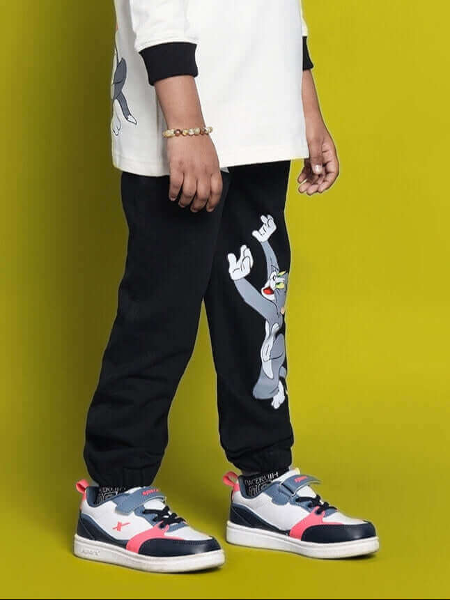 Child wearing black joggers with Tom & Jerry design, standing against a yellow background.