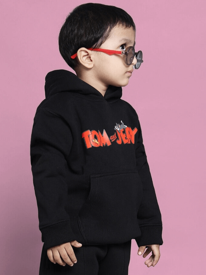Child wearing a black Tom and Jerry oversized hoodie with red lettering, standing against a pink background.
