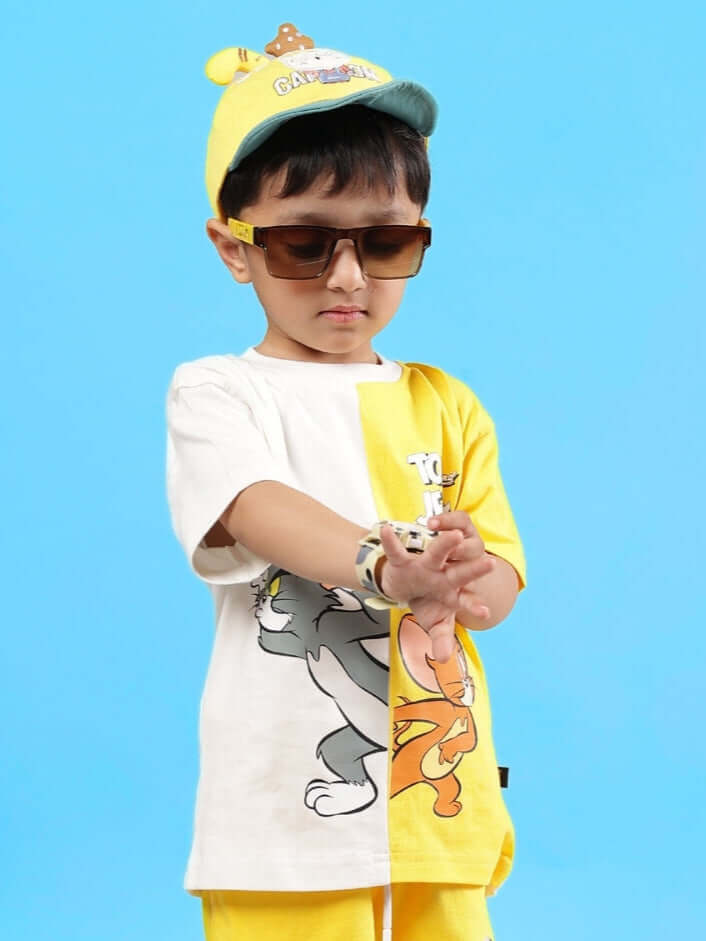 Young boy wearing Tom & Jerry oversized off white-yellow t-shirt, sunglasses, and a colorful cap against a blue background.