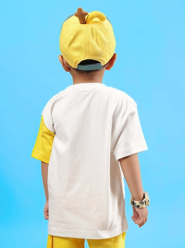 Child wearing Tom & Jerry oversized off-white and yellow T-shirt facing away, paired with yellow cap, against blue backdrop.