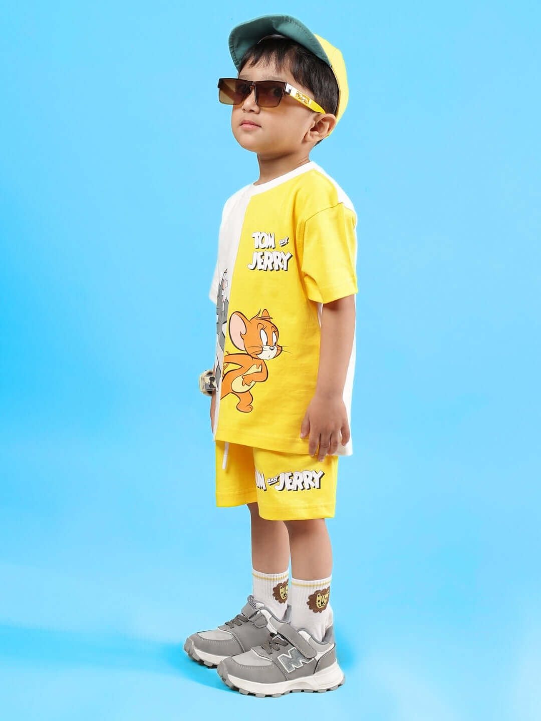 Child wearing yellow Tom & Jerry co-ord set with cap and sunglasses, side view against blue background.