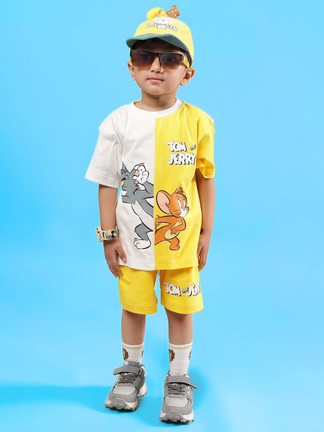 Young boy wearing TOM & JERRY SHORTS FOR BOYS & GIRLS YELLOW with matching shirt and cap, standing against a blue background.