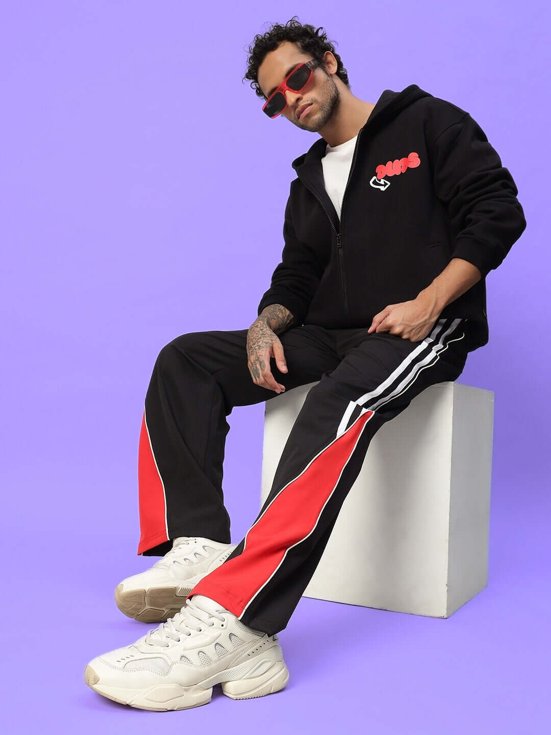 Man in black and red streetwear tracksuit with sunglasses, seated on a white cube against a purple background.