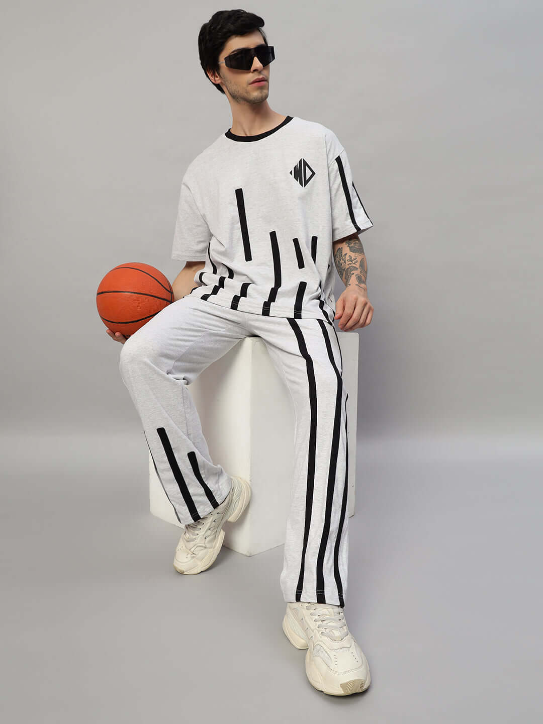 Man in stylish white set with black stripes holding a basketball, sitting on a white block. Trendy athletic fashion in casual setting.