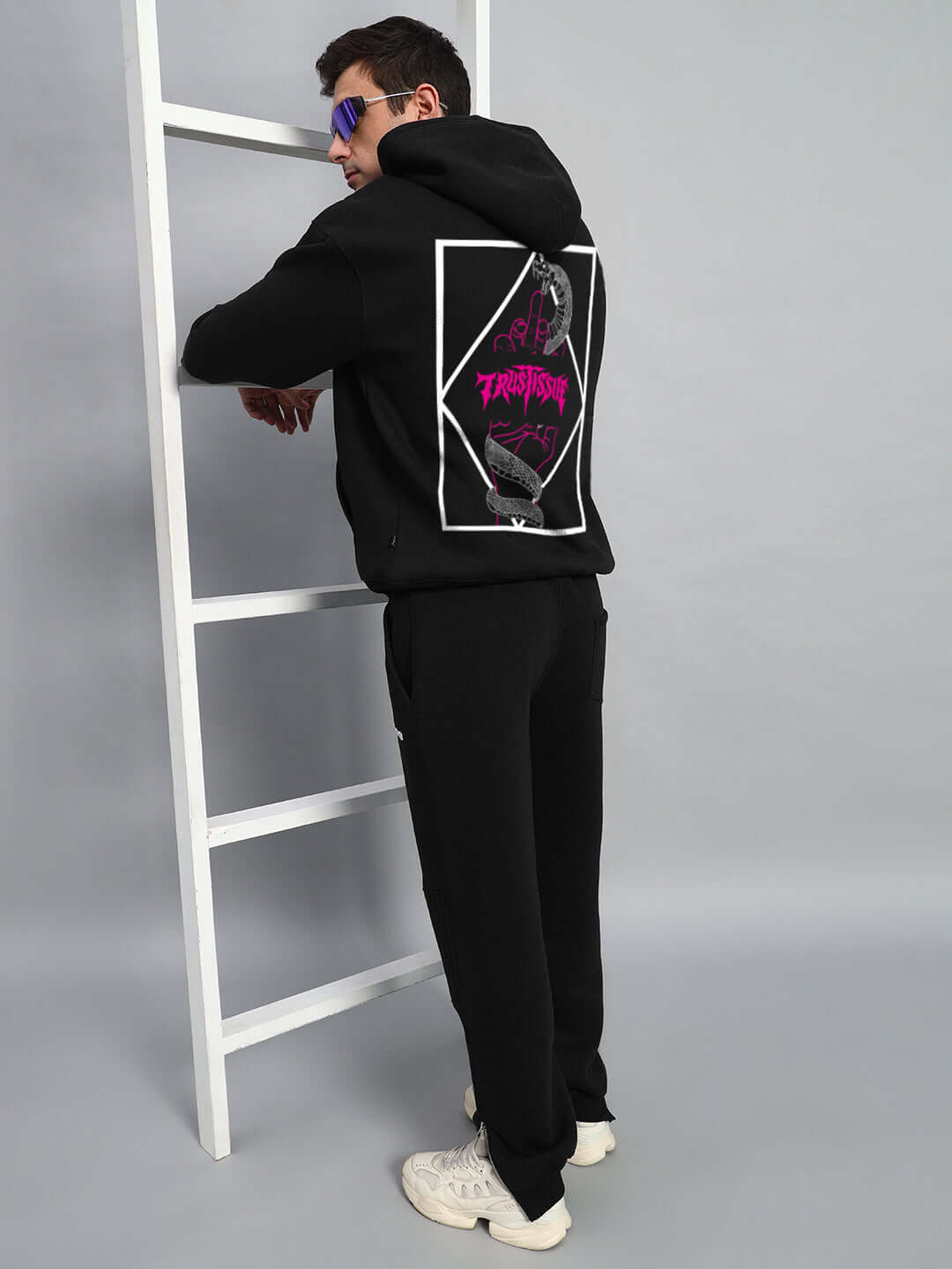 Model wearing Trustissue oversized black hoodie with graphic on back, styled with black pants and white sneakers.