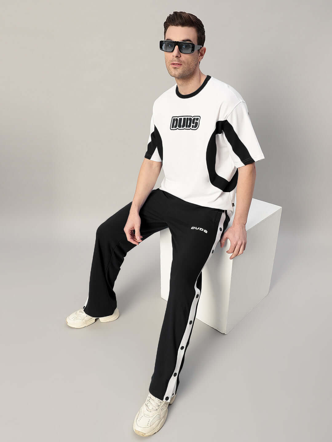 Man wearing EZEL RELAXED FIT CO-ORD SET WHITE-BLACK with sunglasses, sitting on white cube, showcasing comfort and style.