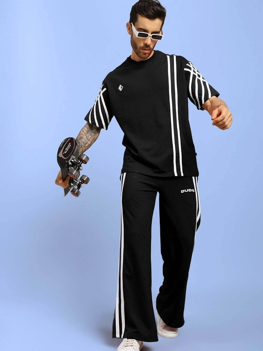 Model wearing black oversized striped t-shirt, holding a skateboard, styled with matching pants, blue background.