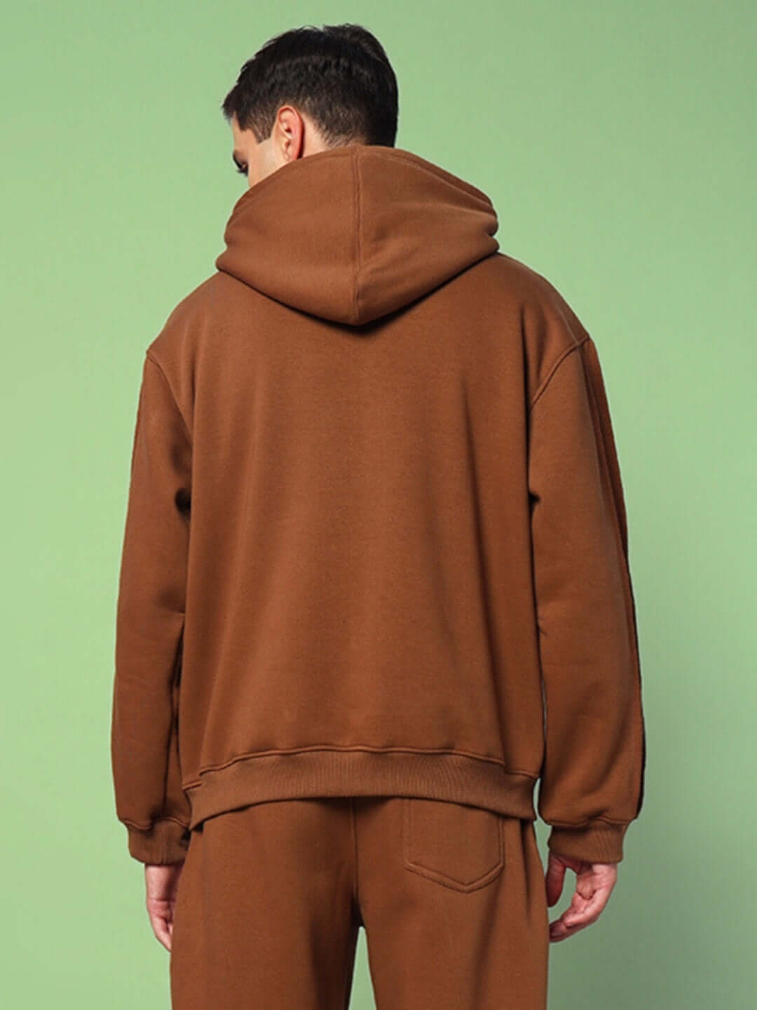 Back view of person wearing URBAN SOLID PLATED FLEECE HOODIE BROWN with hood up and relaxed fit against a green background.