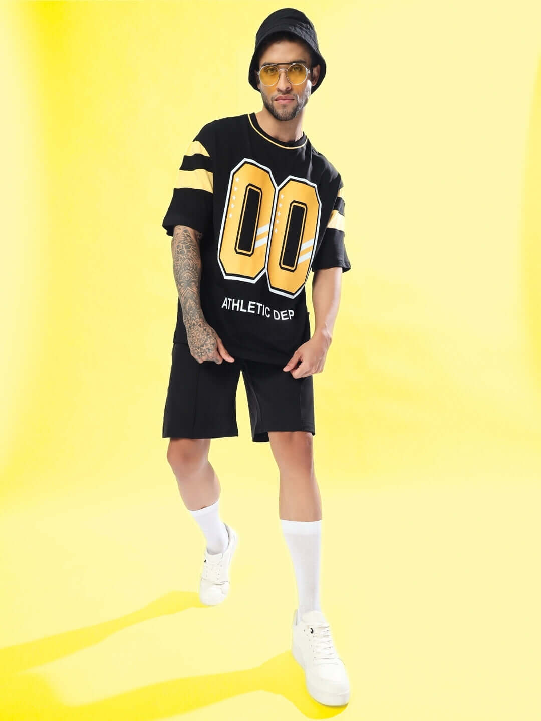 Person wearing sturdy front plated black shorts paired with a sporty black jersey and white sneakers against a vibrant yellow background.