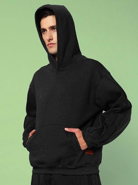 Man wearing URBAN SOLID PLATED FLEECE HOODIE BLACK with hands in pockets, showcasing modern urban style on a green background.