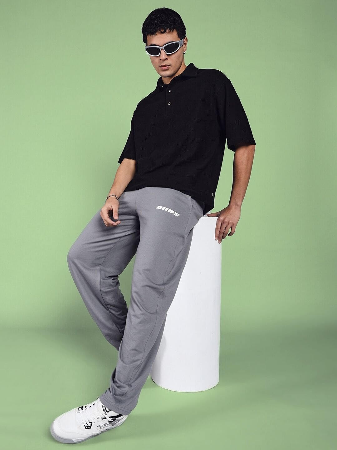 Model wearing Vanilla Relaxed-Fit Joggers in grey, styled with a black polo shirt and sunglasses, against a green backdrop.