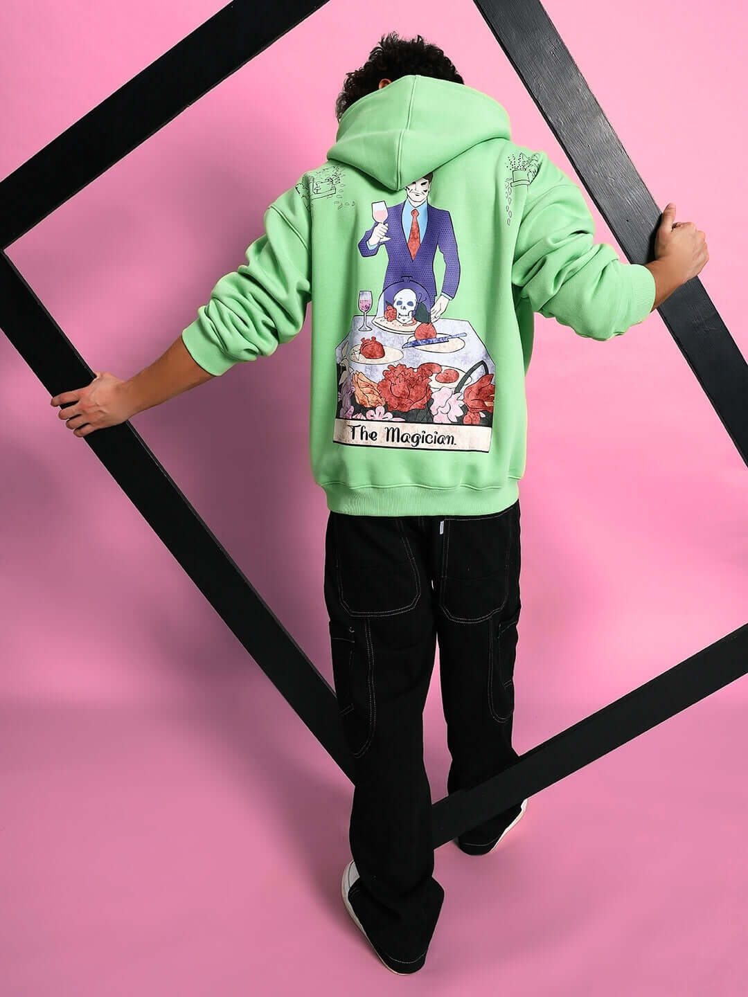 Model wearing VOODO OVERSIZED FLEECE HOODIE GREEN with graphic design on the back, showcasing bold streetwear fashion against a pink background.