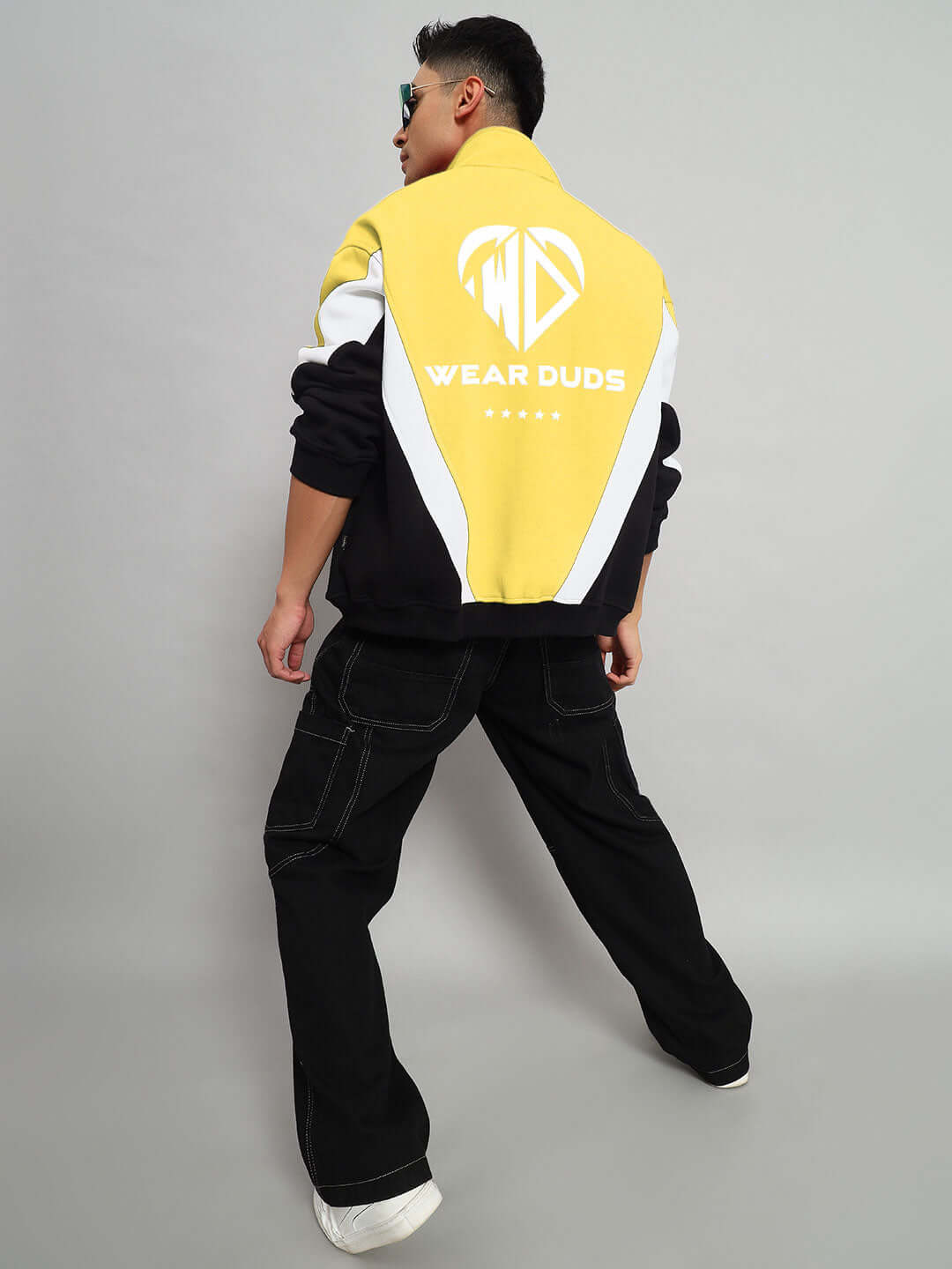 Back view of model wearing yellow WD ALFA Fleece Colorblock Jacket by Wear Duds, featuring bold design and logo, black pants, grey backdrop.