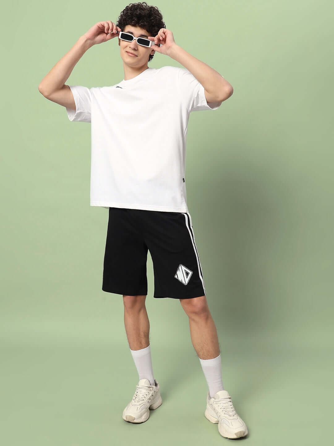 Man wearing WD Premium Regular Fit Black Shorts with white shirt and sunglasses, posing stylishly against a green background.