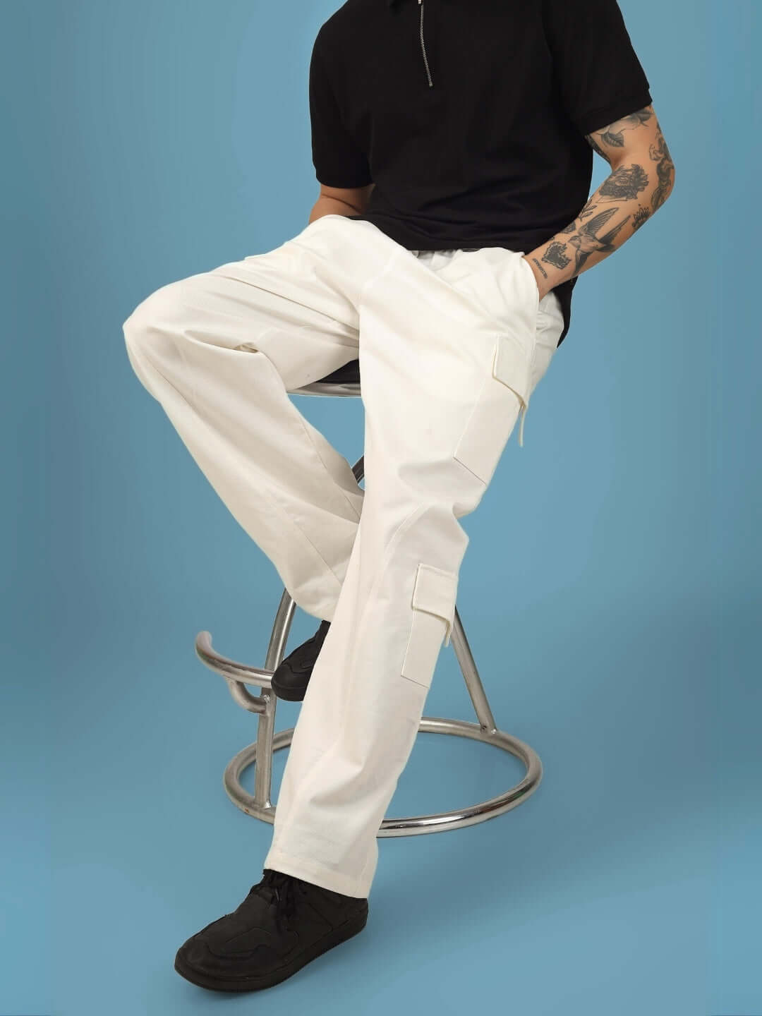 Model wearing 6 pocket white cotton twill cargo pants seated on a stool against a blue background, showcasing casual style.