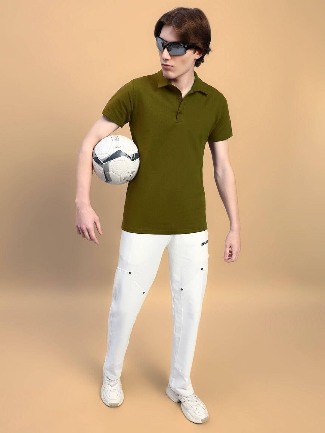 Man in olive green WIDEL regular fit t-shirt holding soccer ball, wearing white pants and sunglasses, standing against beige background