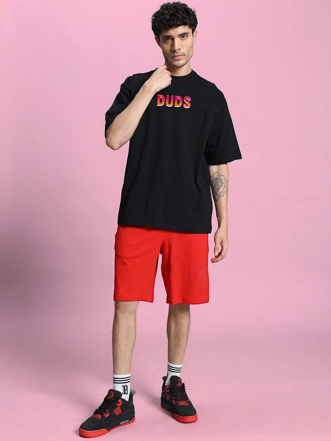 Man wearing WILD CITY OVERSIZED T-SHIRT BLACK with red shorts, standing on pink background