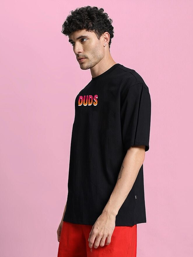 Man wearing WILD CITY OVERSIZED T-SHIRT BLACK with graphic print, styled with red shorts against a pink background.