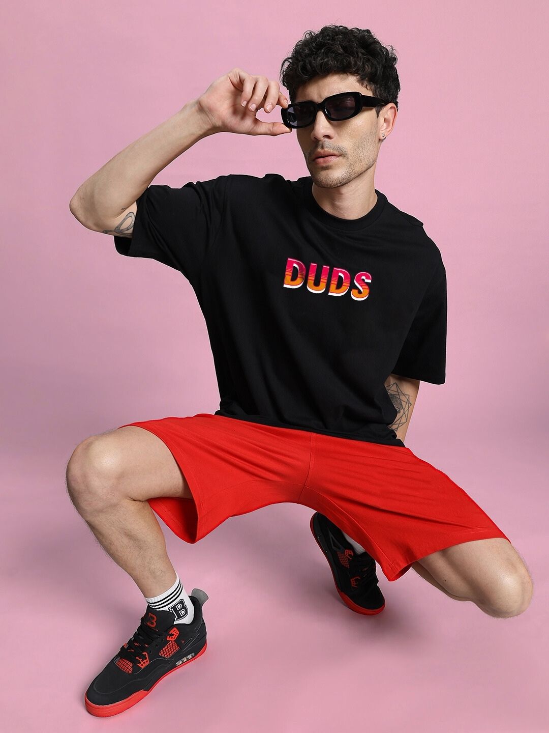Model wearing WILD CITY OVERSIZED T-SHIRT BLACK with red shorts, posing against a pink background, showcasing street fashion style.