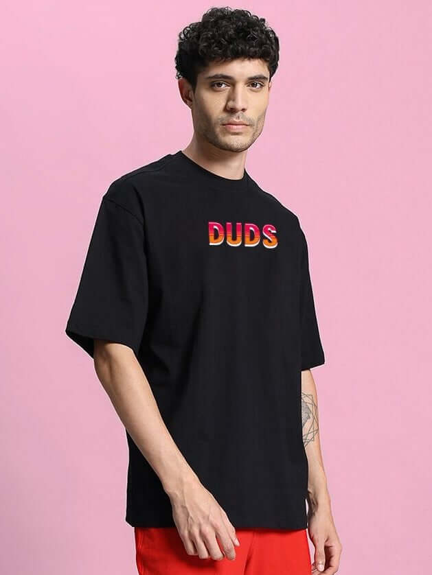 Man wearing WILD CITY OVERSIZED T-SHIRT BLACK with graphic design on pink background.