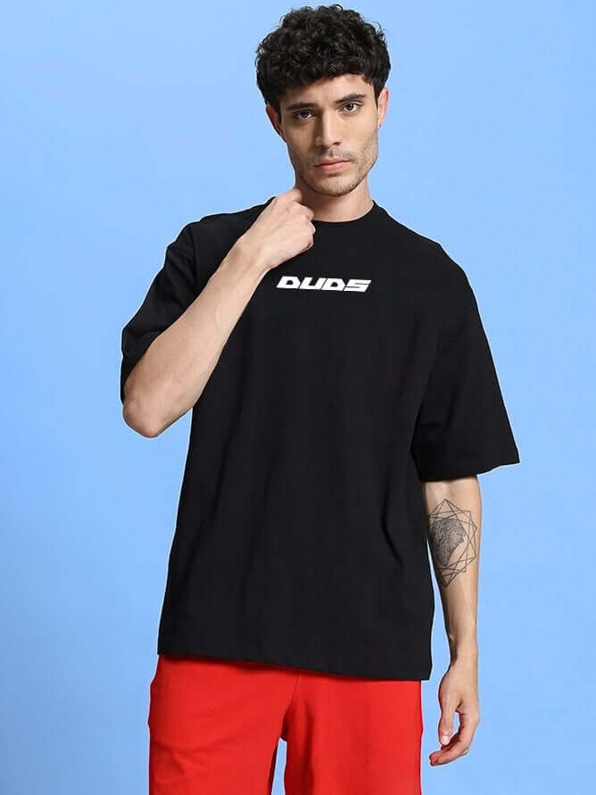 Man wearing WINGED WARRIOR PRINTED OVERSIZED T-SHIRT BLACK with red pants against a blue background.