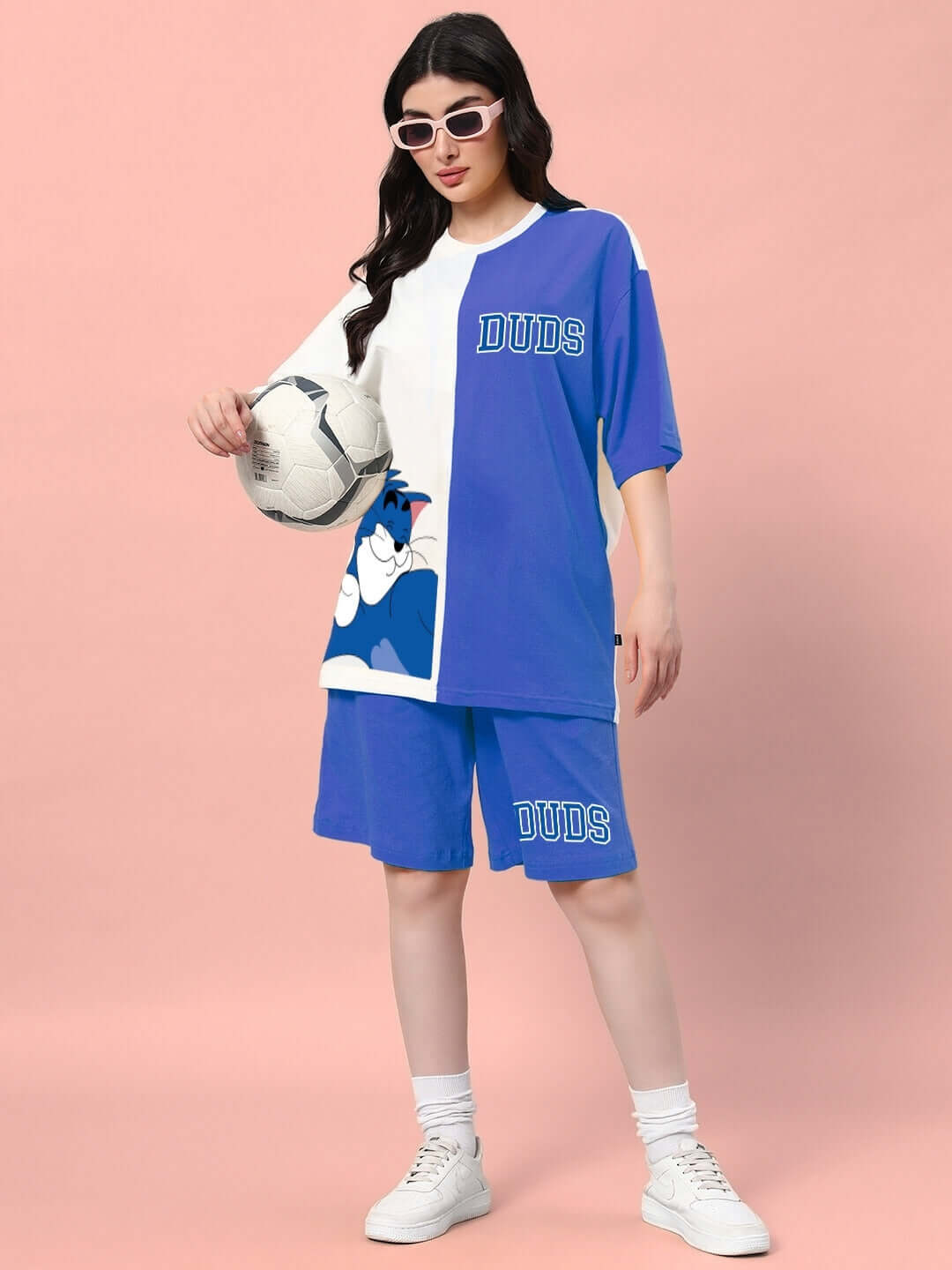 Model wearing DUDS Women's Caper Tom Summer Shorts in blue with matching shirt, holding a soccer ball against a pink background.
