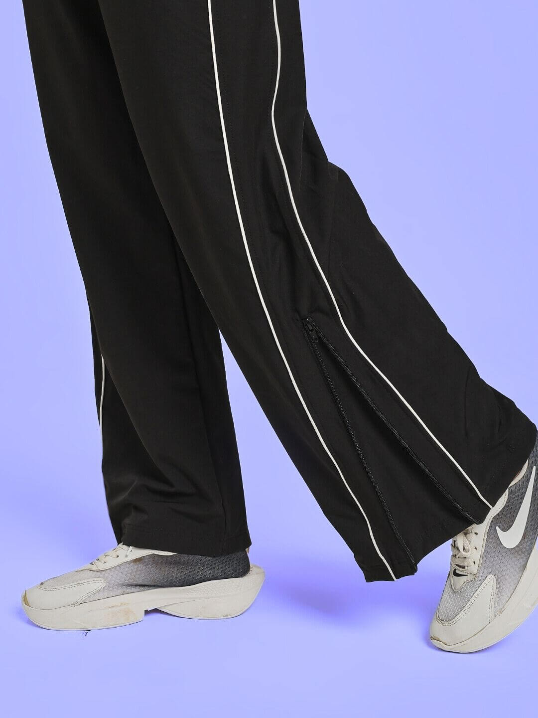 Close-up of WOMEN RELIGHT RELAXED FIT CO-ORD SET BLACK pants with white piping, paired with gray sneakers on a lilac background.