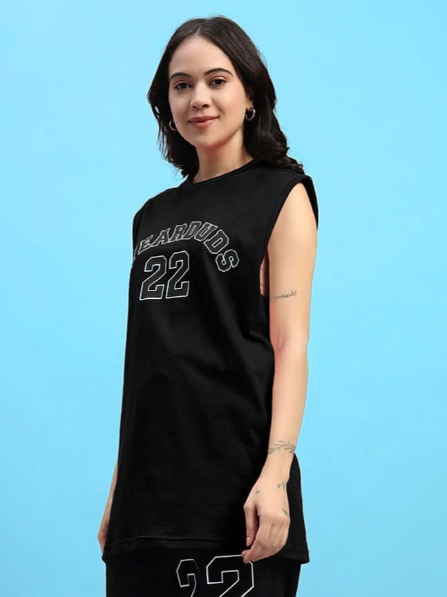 Woman wearing Women's Amiga Gym T-Shirt in black, featuring a casual sleeveless design with "EAROUDS 22" print, standing against blue background.