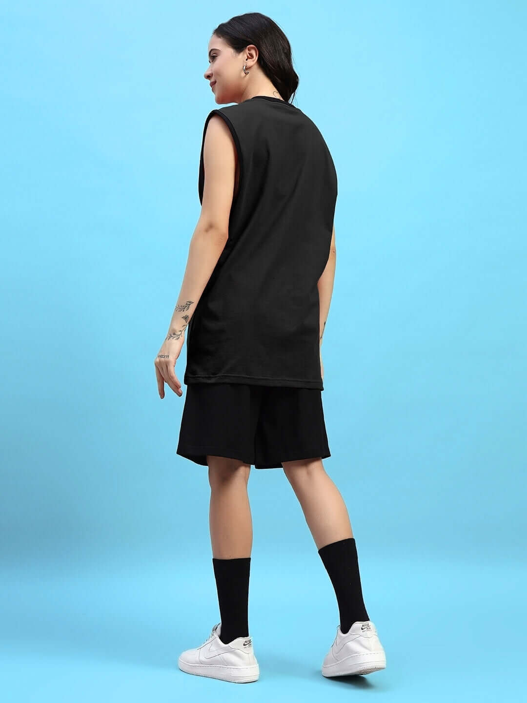 Woman wearing black regular fit shorts and top with sneakers, standing against a blue background.