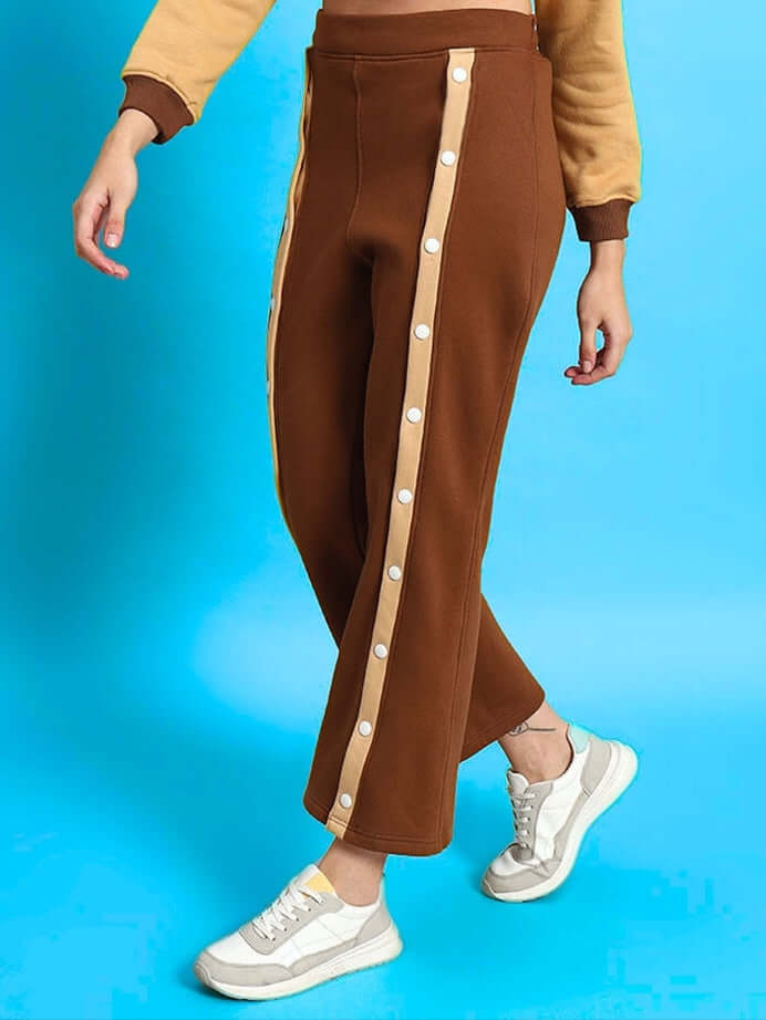 Woman wearing beige-brown aurora fleece front open pants with side buttons, styled with sneakers against a blue background.