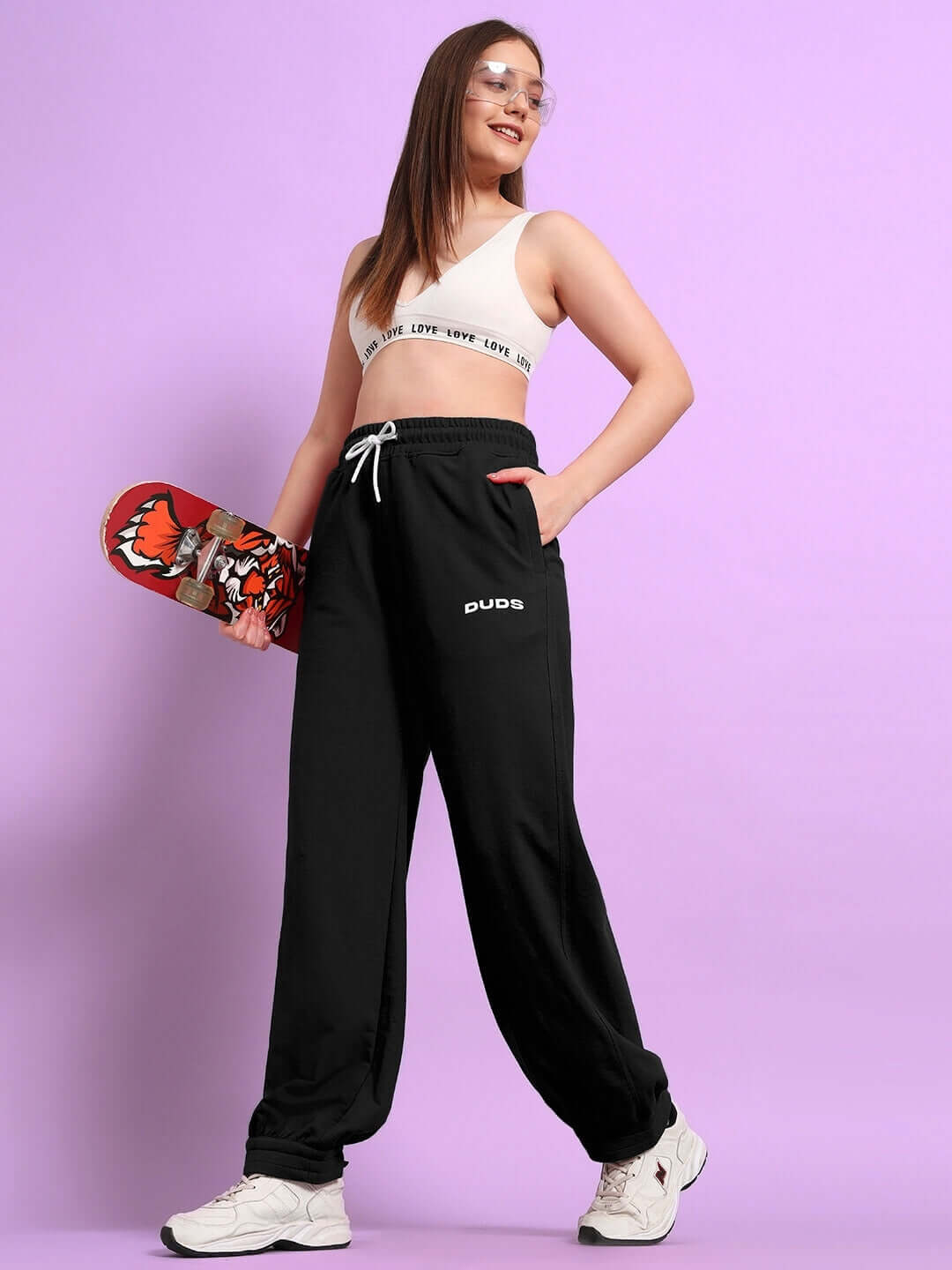 Woman wearing WOMENS AUSTRIAN RELAXED-FIT JOGGERS BLACK holding skateboard on purple background