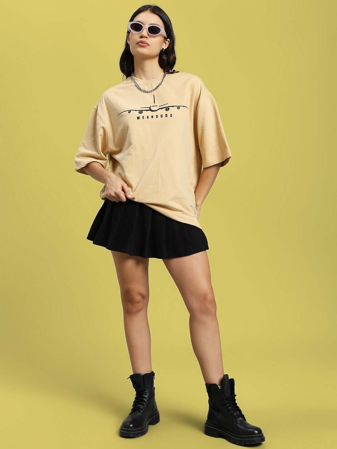 Woman wearing oversized beige aviator designer T-shirt with black skirt, styled with boots and sunglasses, against a yellow background.